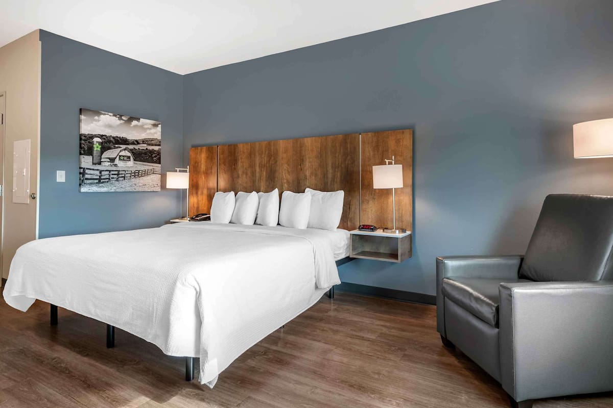Une chambre d’hôtel avec un grand lit, une tête de lit en bois, deux tables de chevet avec des lampes, un fauteuil gris et une œuvre d’art accrochée au mur.