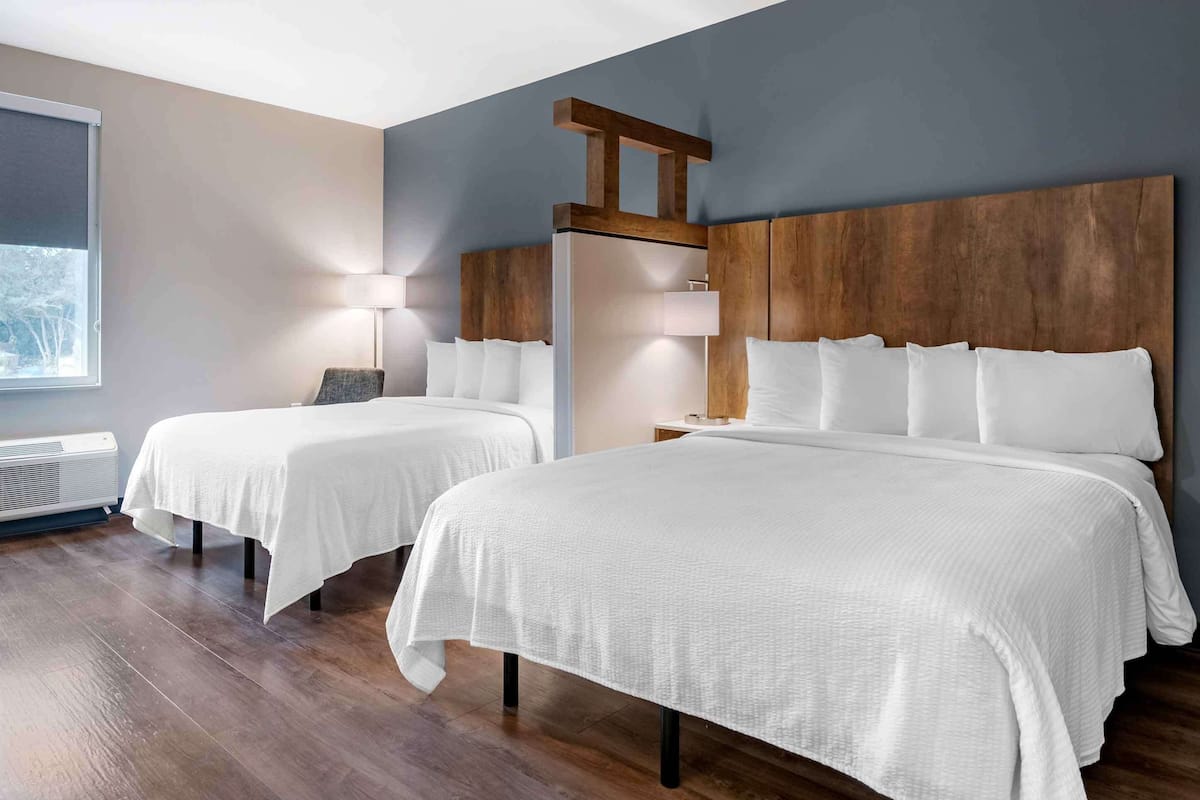 Une chambre d’hôtel avec deux lits, une tête de lit en bois, une table de chevet avec une lampe et une fenêtre avec des stores.