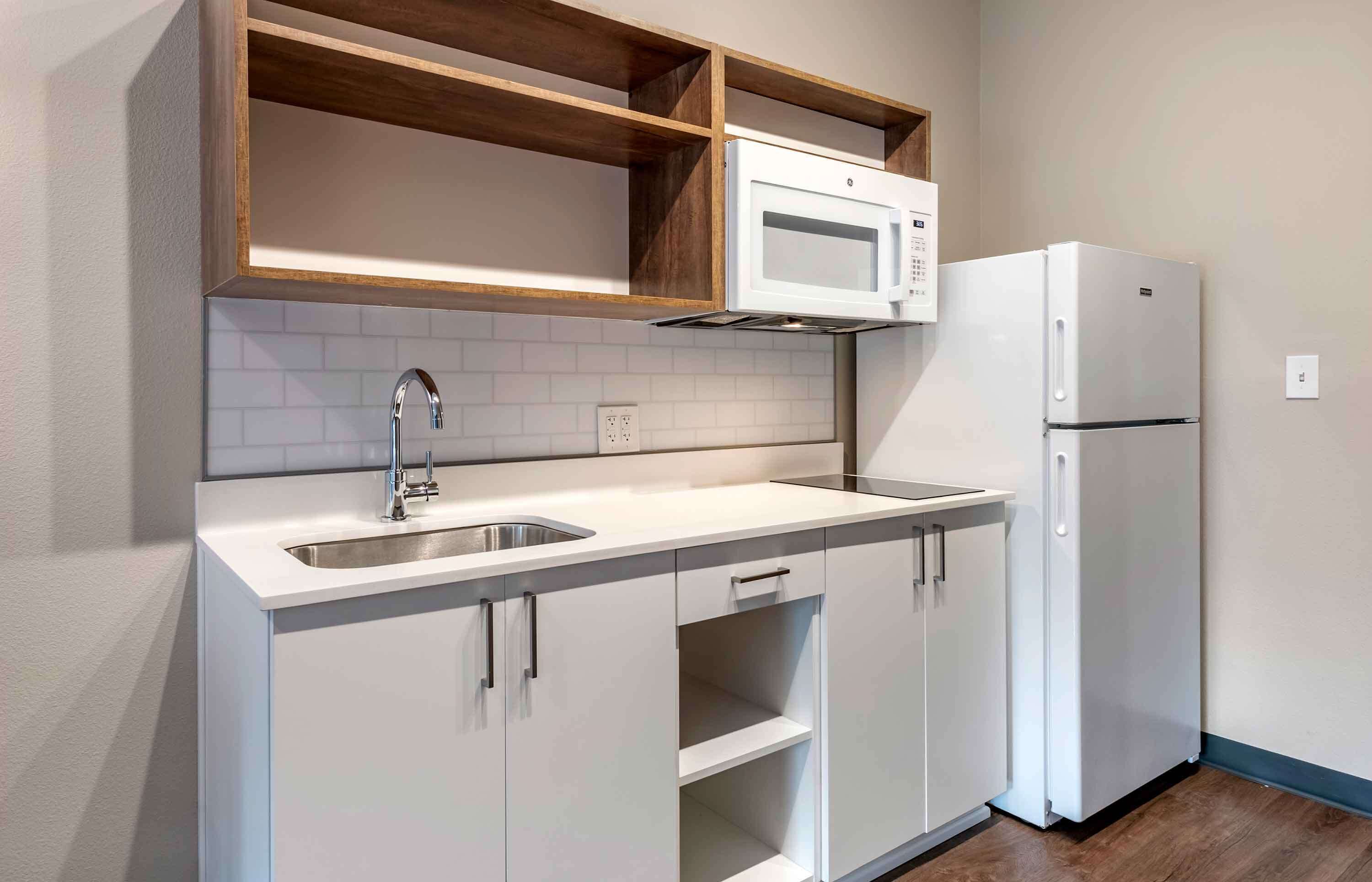 A compact kitchen with a built-in microwave, a refrigerator, a sink with a faucet, and wooden shelves above the countertop.