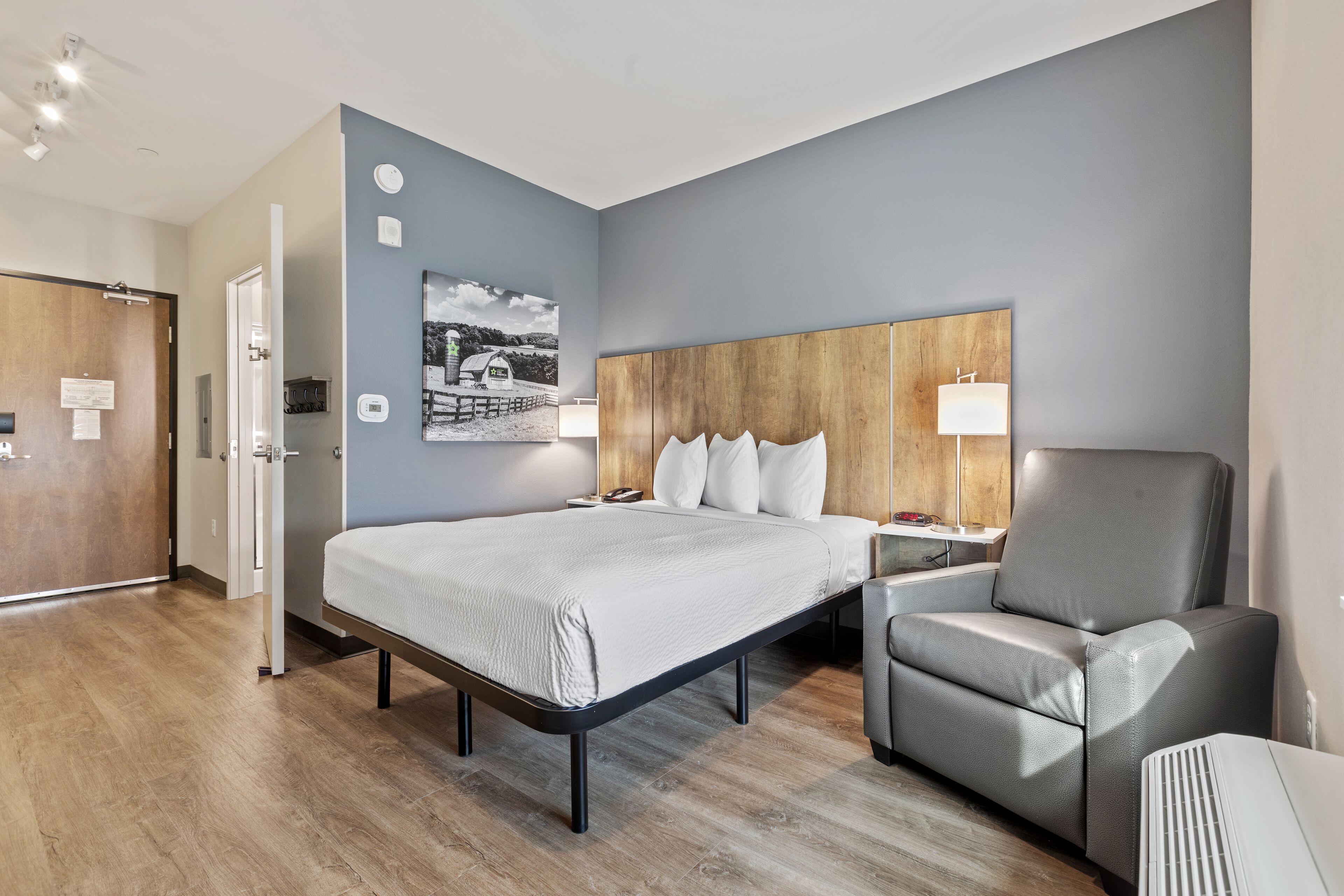 Une chambre d’hôtel moderne avec un grand lit, un fauteuil gris, une tête de lit en bois, un téléviseur fixé au mur et un tableau représentant une scène de plage.