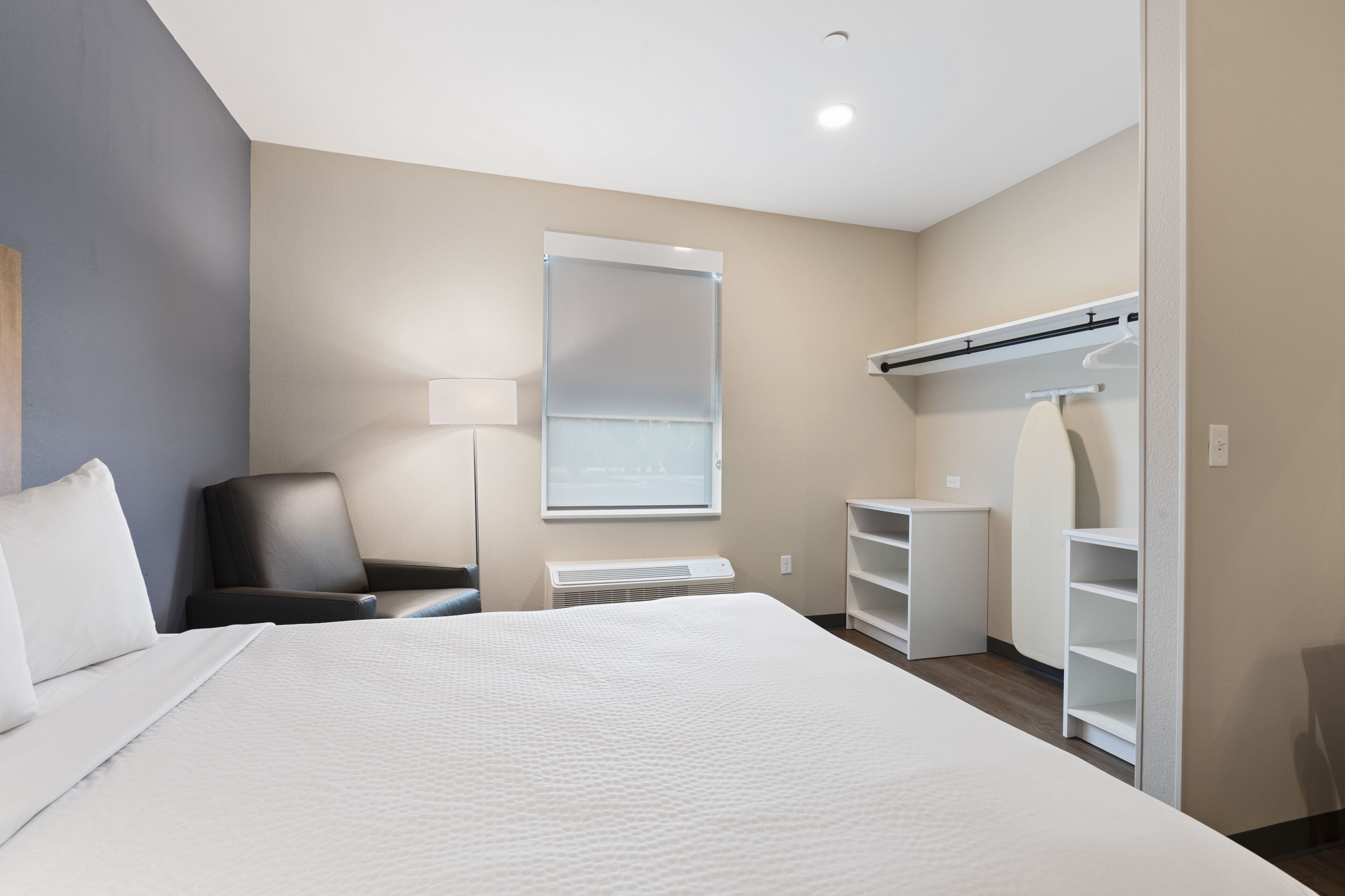 A hotel room with a large bed, a desk, a chair, a wardrobe, and a window with blinds.