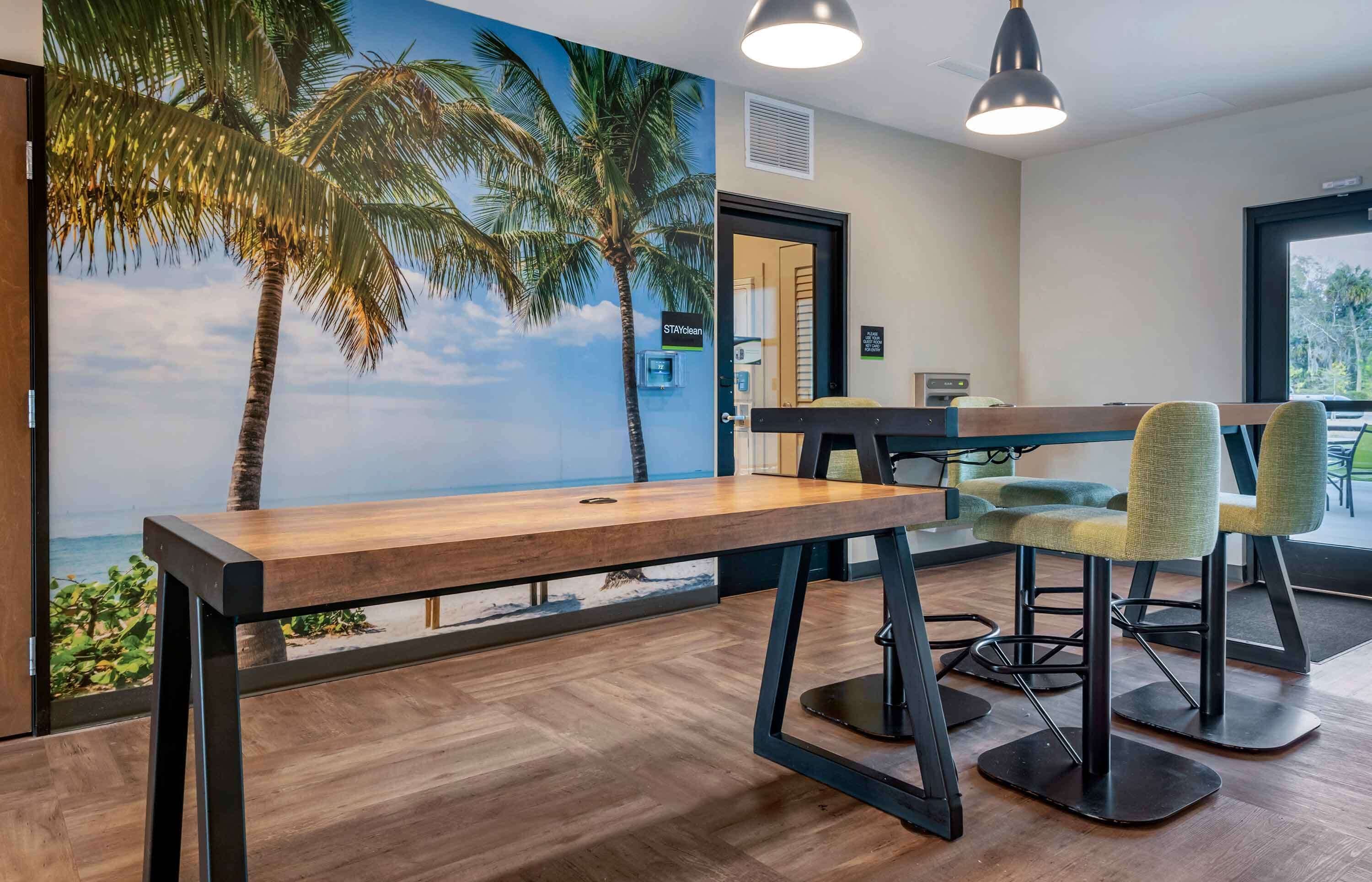 Une pièce avec une table et des chaises en bois, une fresque représentant une scène de plage et une vue sur l’extérieur.