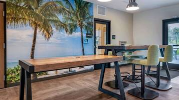 Une pièce avec une table et des chaises en bois, une fresque représentant une scène de plage et une vue sur l’extérieur.