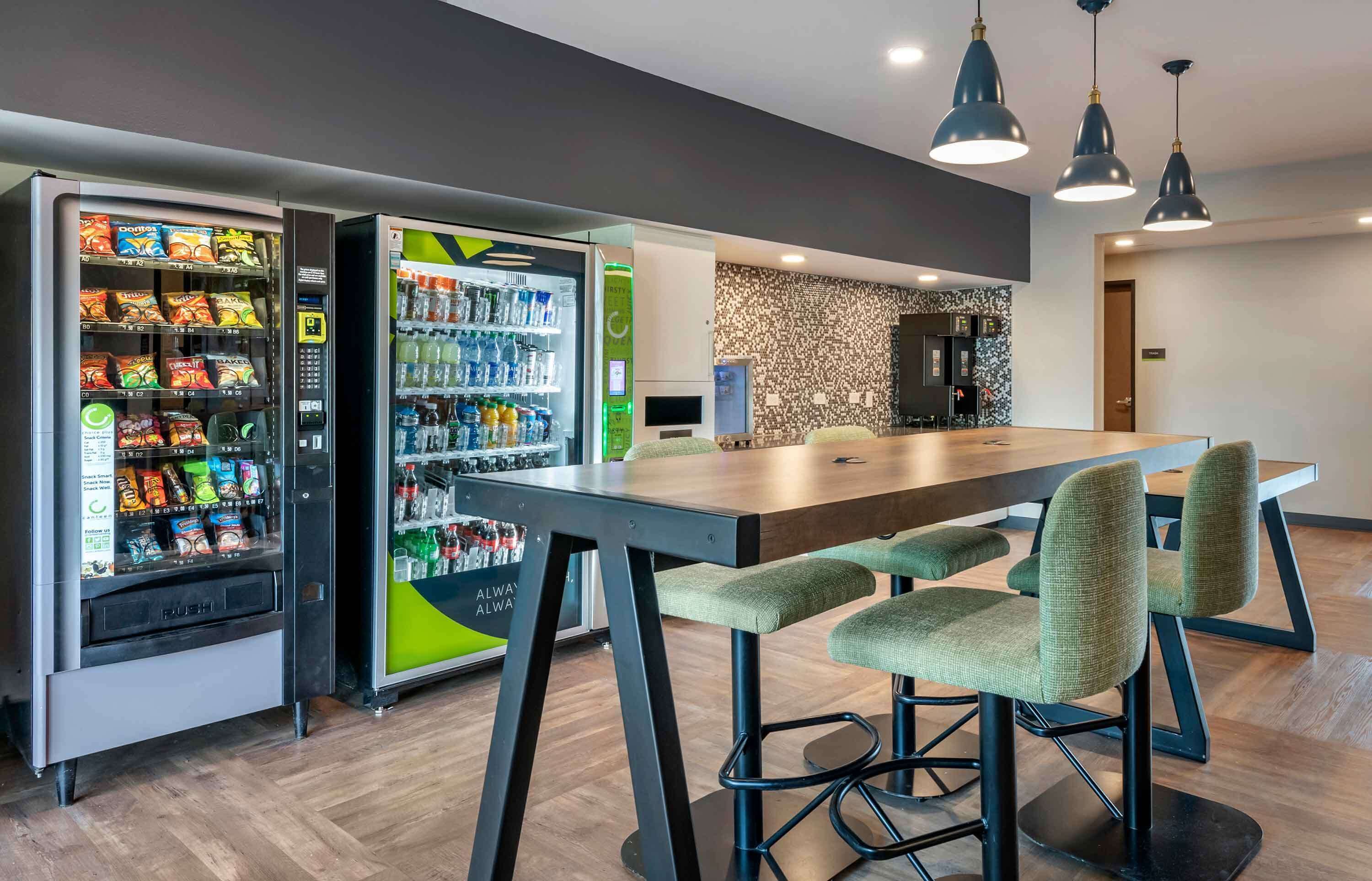 A modern interior with vending machines, a wooden table, and green cushioned chairs.