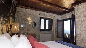 Ein Schlafzimmer mit Steinwand, Holzboden, einem Bett mit weißer und roter Bettwäsche, einem Nachttisch mit Lampe, einem Spiegel und einem Fenster mit Blick aufs Meer.