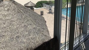 A beach view from a window with a thatched roof, a pool, and a sandy beach. - Oceanfront condo, beach, pool. 4 eateries on premises. Rent kayaks and jet skis. (Fort Lauderdale)