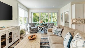 Un salon spacieux doté d’une grande fenêtre, d’une table basse en bois, d’un canapé beige, de fauteuils à motifs et d’un coin cuisine avec des meubles blancs.