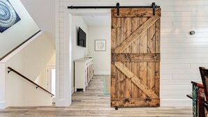 Un couloir doté d’une porte coulissante en bois de style grange, d’un escalier avec une rampe et d’une vue sur une pièce où se trouvent un téléviseur et un tableau encadré au mur.