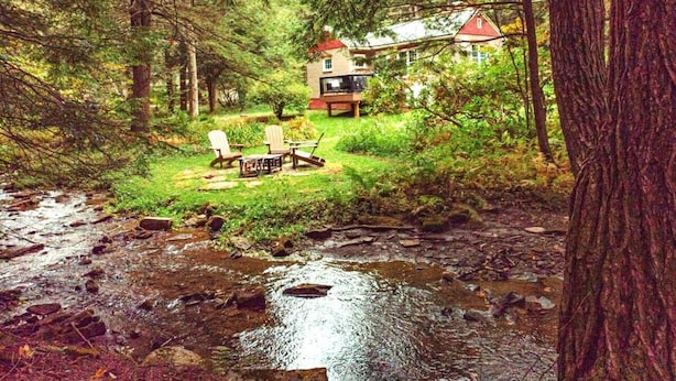 A cabin with a red roof surrounded by a forest, a stream in the foreground, and outdoor furniture. - Romantic Waterfront Cottage for Cherry Springs (Coudersport)
