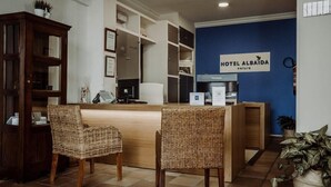 A reception area with a wooden desk, two wicker chairs, a blue wall with a sign reading "HOTEL ALBAIDA nature", and a plant in the corner.
