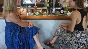 Two women seated at a bar with a coffee machine and various bottles in the background.