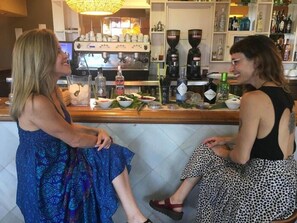 Zwei Frauen sitzen an einer Bar, im Hintergrund eine Kaffeemaschine und verschiedene Flaschen.