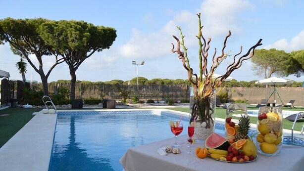 Zona de piscina con una mesa dispuesta con frutas y bebidas, rodeada de árboles y un cielo despejado.