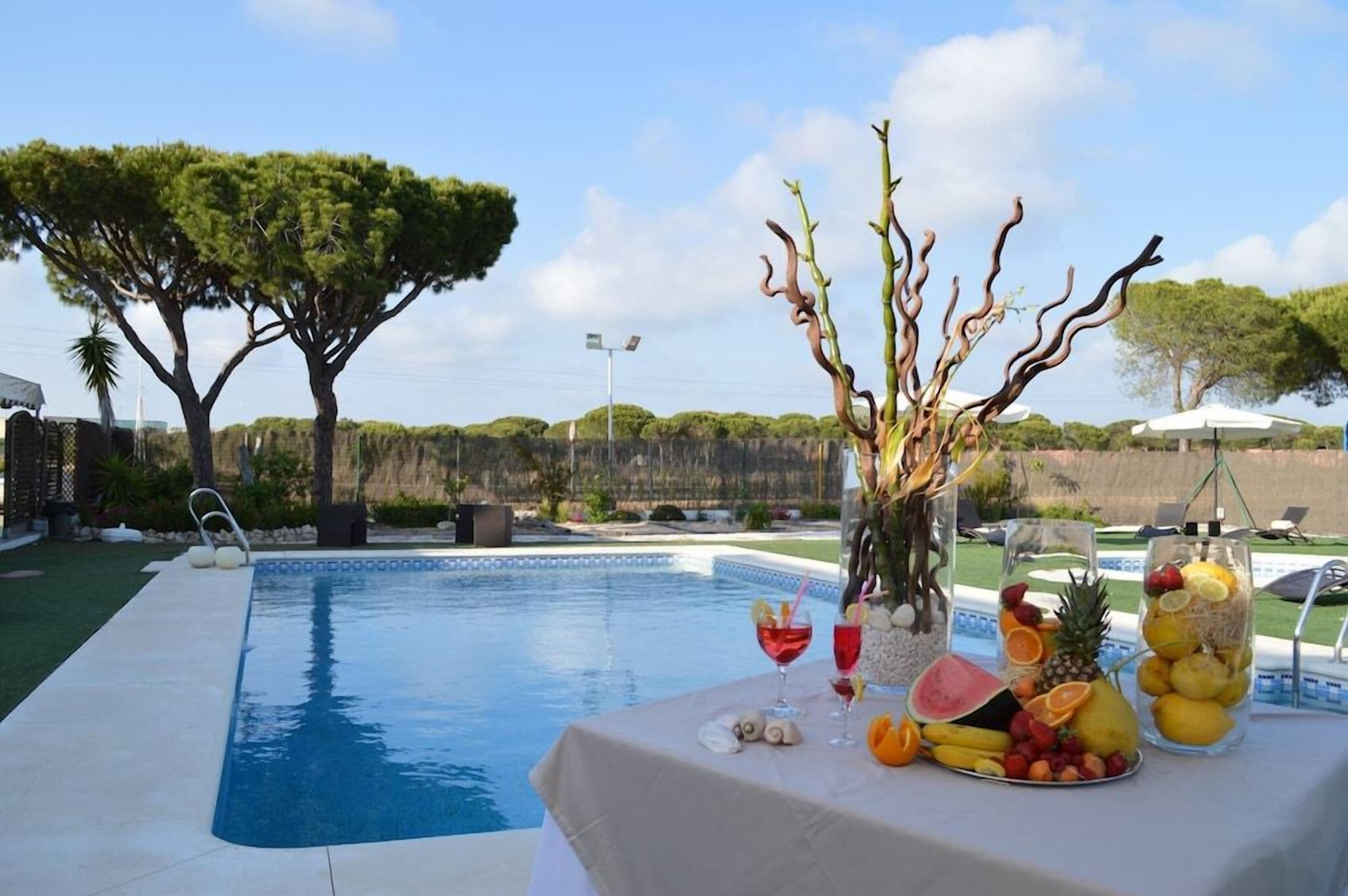 A poolside area with a table set with fruit and drinks, surrounded by trees and a clear sky.