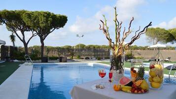 A poolside area with a table set with fruit and drinks, surrounded by trees and a clear sky.