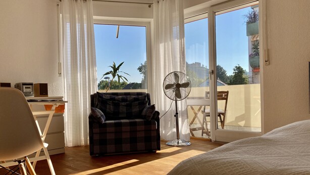 Una habitación con cama, una silla, un escritorio, un ventilador y vistas al exterior.