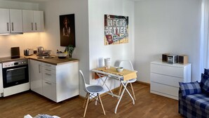 Cocina moderna con armarios blancos, horno empotrado y zona de comedor con una mesa y sillas pequeñas. Hay un cuadro en la pared y una ventana con cortinas.
