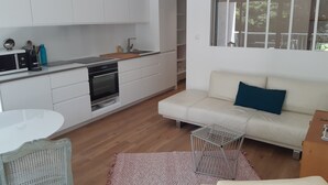 Une cuisine moderne avec des armoires blanches, un four encastré et un évier. On y trouve un canapé blanc avec un coussin bleu et une petite table à structure métallique.