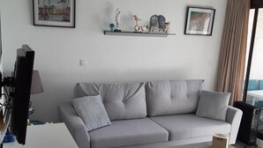 Una sala de estar moderna con un sofá gris, una mesa de centro y un televisor sobre un mueble bajo. Hay objetos decorativos en un estante sobre el sofá y una ventana con cortinas.