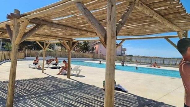 Una pérgola de madera sobre un área de piscina con sillones de descanso y gente relajándose.