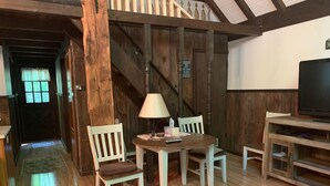 Una habitación rústica con paredes de madera y una escalera. Hay una mesa redonda con sillas, una lámpara y un televisor sobre un soporte de madera.