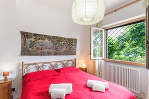 A bedroom with a bed, a painting, a window with a view, a bedside lamp, and a radiator. - GuestHost - Charming and bright flat for 5 people in Cernobbio, in the hamlet of Rovenna, on the way up to mount Bisbino.The apartment, furnished with vintage and modern furniture, comes with private parking and enjoyable outdoor spaces, like the pergola (Cernobbio)