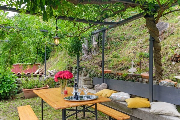A wooden table with benches, a vase with flowers, and a lantern under a pergola in a garden setting. - GuestHost - Charming and bright flat for 5 people in Cernobbio, in the hamlet of Rovenna, on the way up to mount Bisbino.The apartment, furnished with vintage and modern furniture, comes with private parking and enjoyable outdoor spaces, like the pergola (Cernobbio)