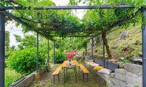A pergola with a table and benches, surrounded by greenery and stone walls. - GuestHost - Charming and bright flat for 5 people in Cernobbio, in the hamlet of Rovenna, on the way up to mount Bisbino.The apartment, furnished with vintage and modern furniture, comes with private parking and enjoyable outdoor spaces, like the pergola (Cernobbio)