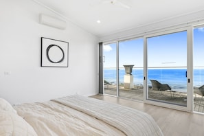 A bedroom with a large bed, a ceiling fan, a framed artwork, and a sliding glass door leading to a balcony with a view of the sea.