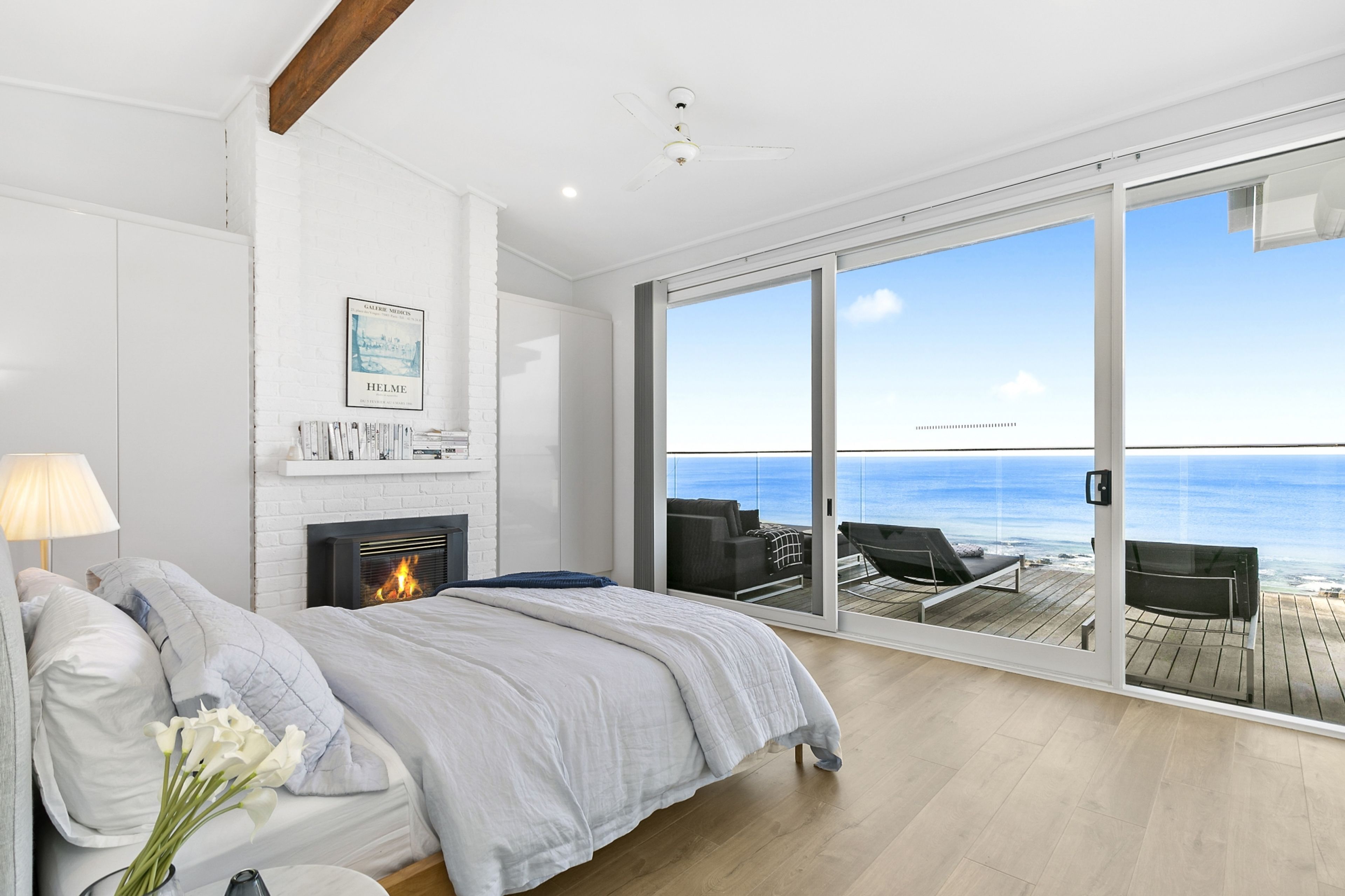 A bedroom with a bed, a fireplace, a view of the ocean, and a balcony with outdoor furniture.
