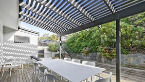 Een buitenpatio met een witte tafel en stoelen, een pergola erboven en een stenen muur begroeid met groen.