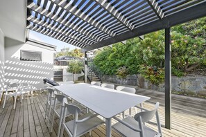 Une terrasse extérieure avec une table et des chaises blanches, une pergola au-dessus et un mur de pierre orné de verdure.