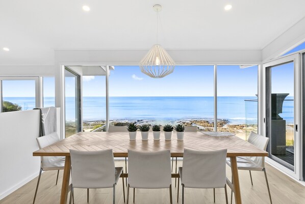 Un coin repas avec vue sur la mer, une table en bois, quatre chaises et un luminaire suspendu.