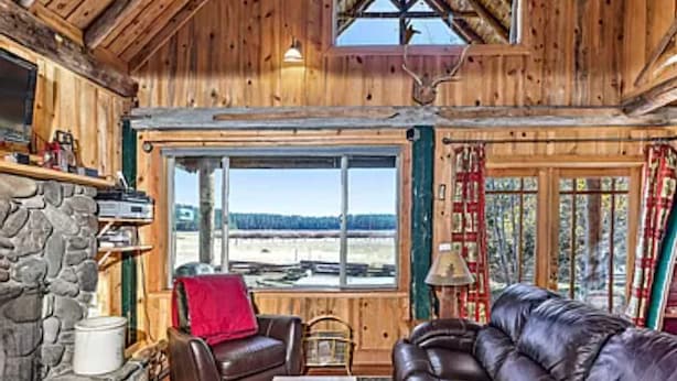 Interior acogedor de una cabaña con chimenea de piedra, sofás de cuero y un amplio ventanal con vistas al exterior.