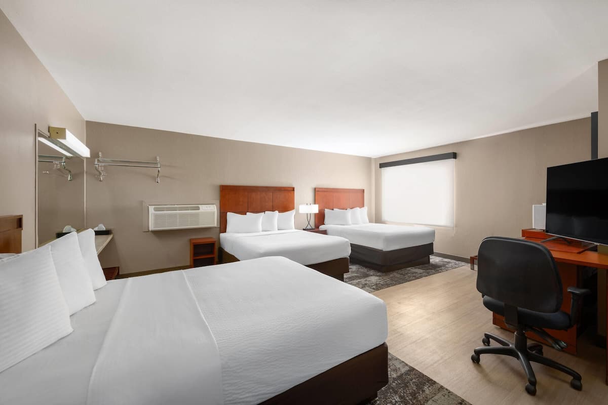Une chambre d’hôtel avec trois lits, un bureau avec une chaise, une télévision et une grande fenêtre.