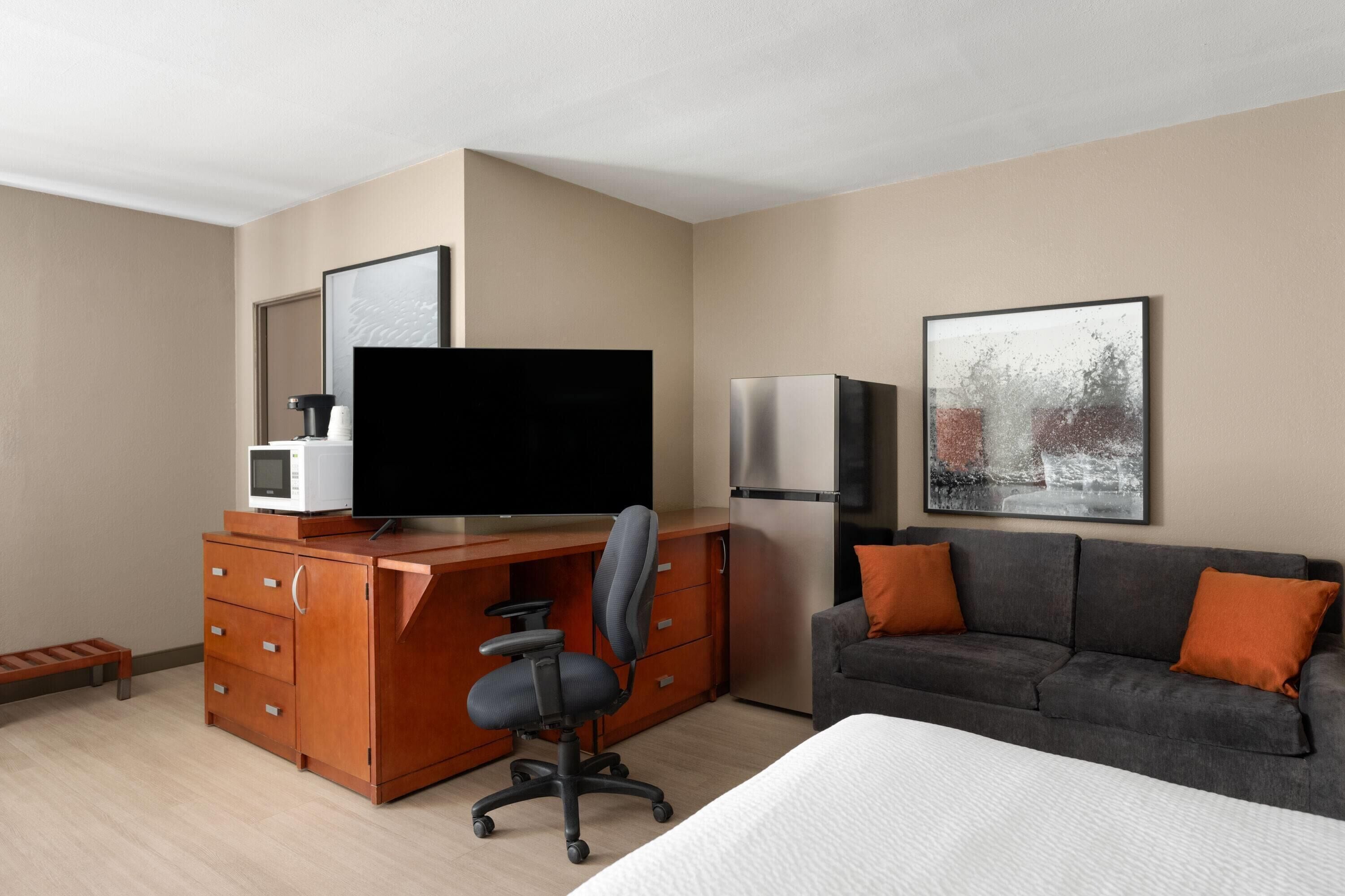 Une chambre d’hôtel moderne avec un bureau en bois, un téléviseur à écran plat, un réfrigérateur, un canapé gris avec des coussins orange et un tableau encadré au mur.