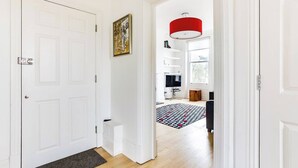 Ein Flur mit einer weißen Tür, einem gerahmten Kunstwerk, einer roten Pendelleuchte und einem gemusterten Teppich, der zu einem Raum mit Fernseher und Fenster führt.