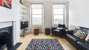 Ein modernes Wohnzimmer mit Kamin, zwei Fenstern, einem schwarzen Ledersofa und einem gemusterten Teppich.