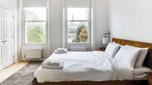 Ein Schlafzimmer mit einem großen Bett, zwei Fenstern mit weißen Jalousien, einem Heizkörper und Blick auf ein Gebäude und Bäume.