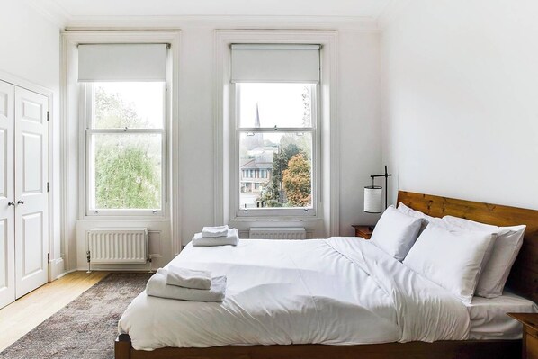 A bedroom with a large bed, two windows with white blinds, a radiator, and a view of a building and trees outside.