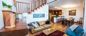 Uma sala de estar ampla com escada de madeira, área de jantar e cozinha ao fundo.