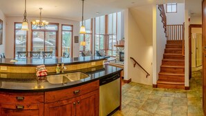 A kitchen with a granite countertop, wooden cabinets, a dishwasher, and a stone fireplace. There is a dining area with a table and chairs, and a staircase leading to another level. - Woodhaven #11 by Bear Country (Sun Peaks)