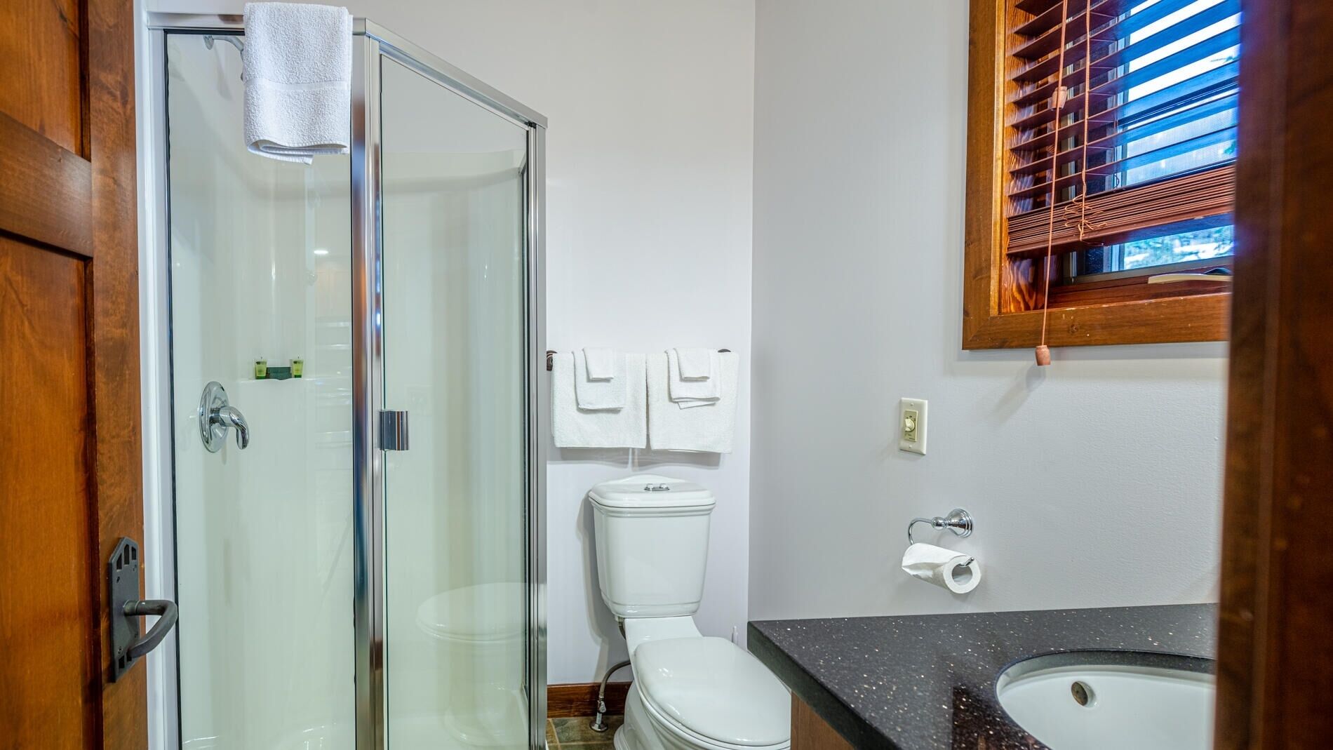 Une salle de bain avec une douche à parois de verre, des toilettes, un lavabo et une fenêtre avec des stores.