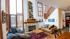 A living room with a stone fireplace, a wooden staircase, and a large window. - Woodhaven #11 by Bear Country (Sun Peaks)