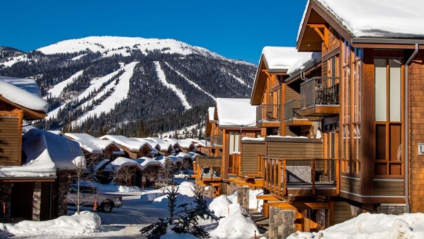 A snow-covered lodge with wooden architecture, a ski slope in the background, and a clear blue sky. - Woodhaven #11 by Bear Country (Sun Peaks)