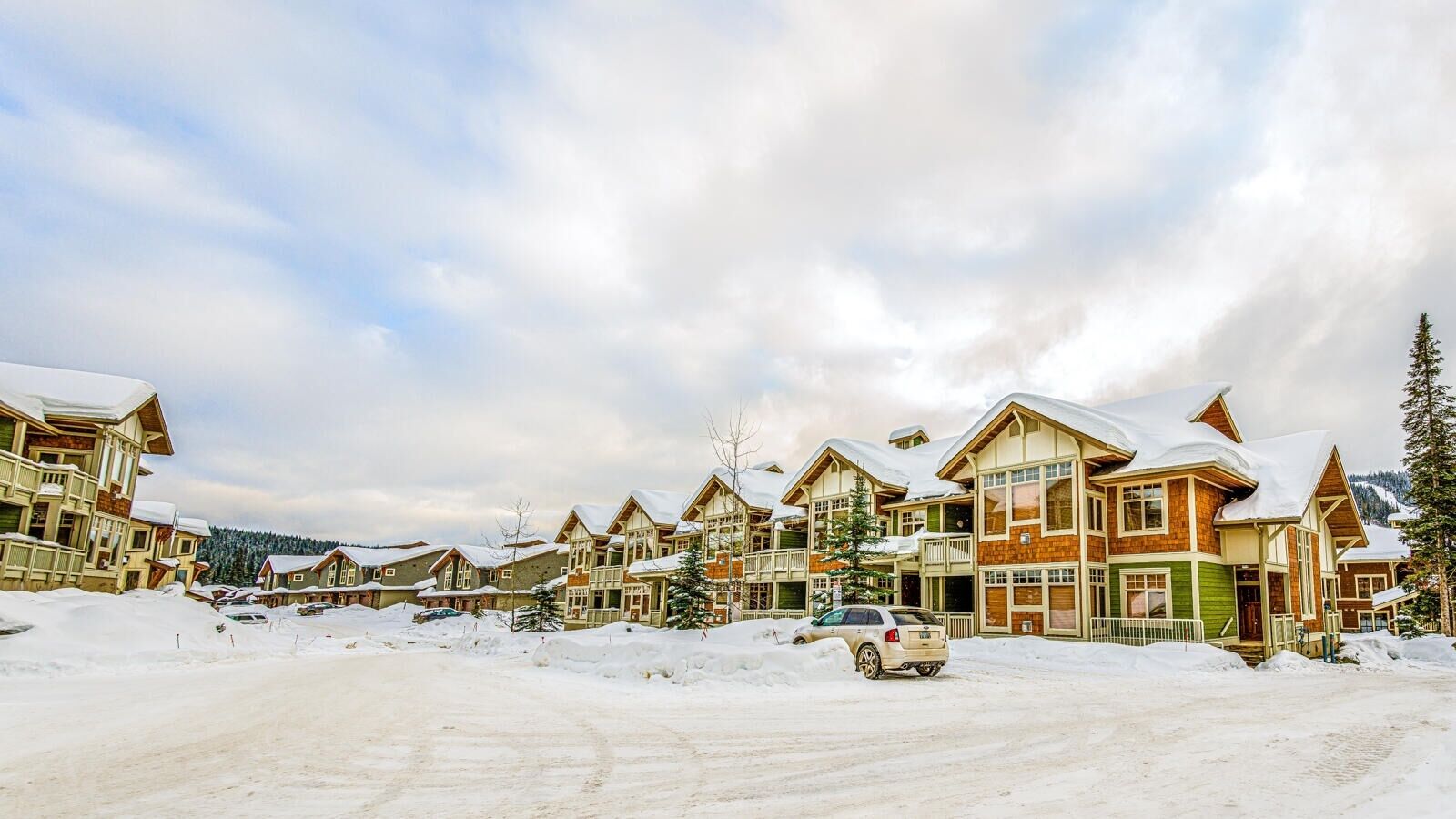 A row of log cabin-style buildings with snow-covered roofs, a parking area, and a mountainous backdrop.