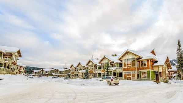 A row of chalet-style buildings with snow-covered roofs, a clear sky, and a snowy ground. - Settlers Crossing #61 by Bear Country (Sun Peaks)