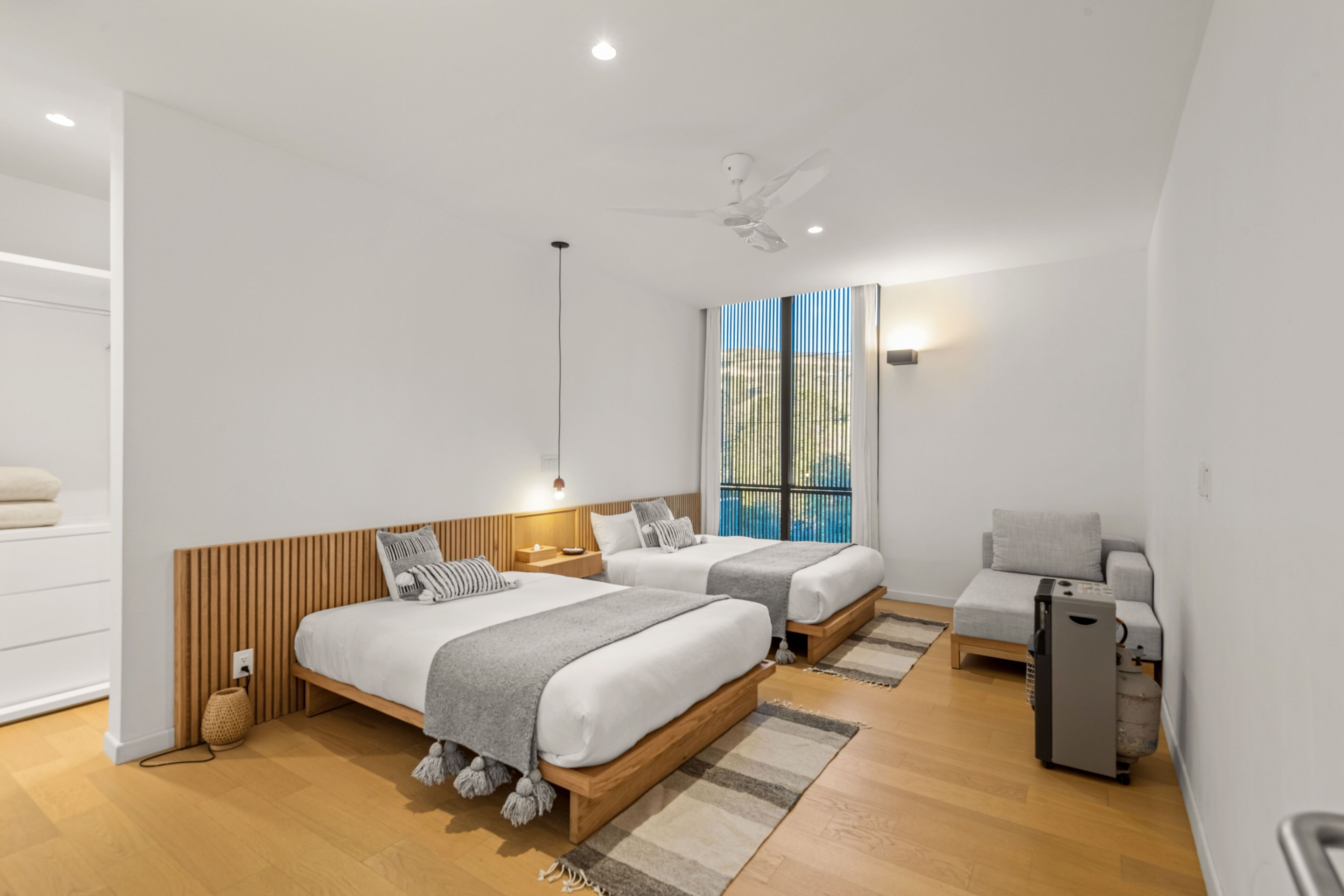 Una habitación de hotel moderna con dos camas, un ventilador de techo, un pequeño sofá, un calentador y una ventana con persianas.
