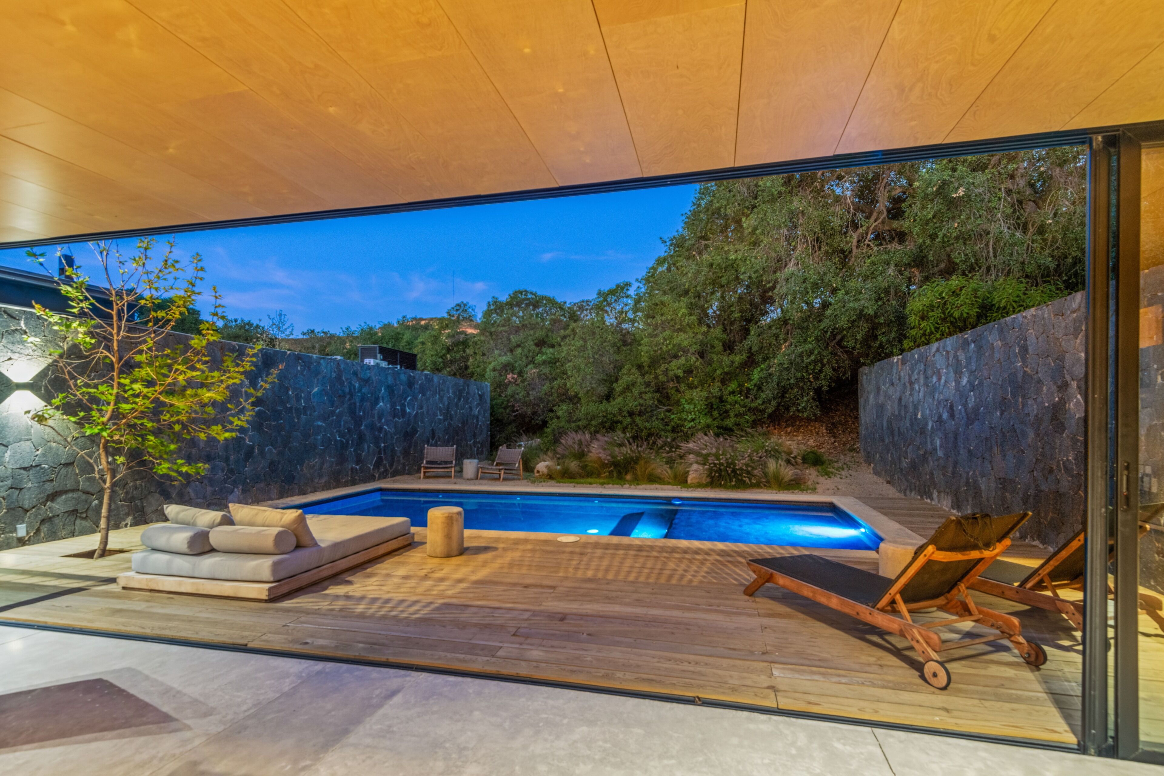A modern outdoor pool area with a wooden deck, lounge chairs, and a stone wall.