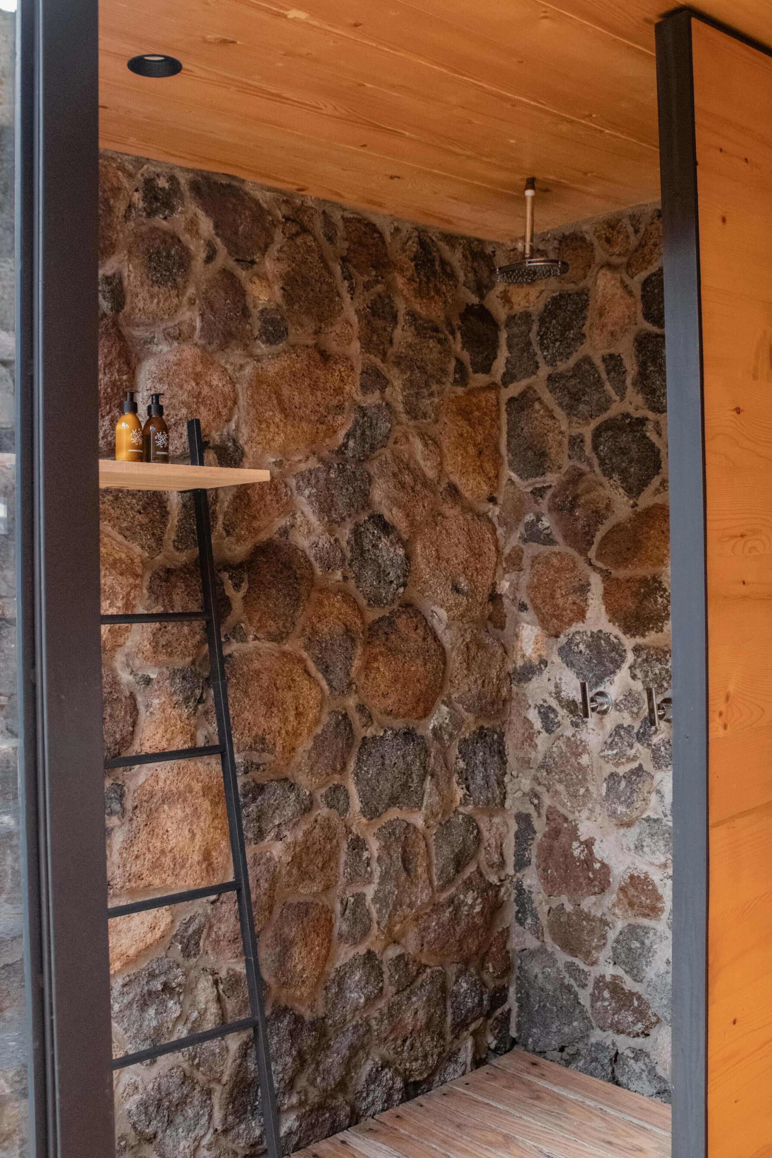 Un espace douche aux murs en pierre, avec une étagère en bois où sont rangés les articles de toilette, une échelle et un pommeau de douche.
